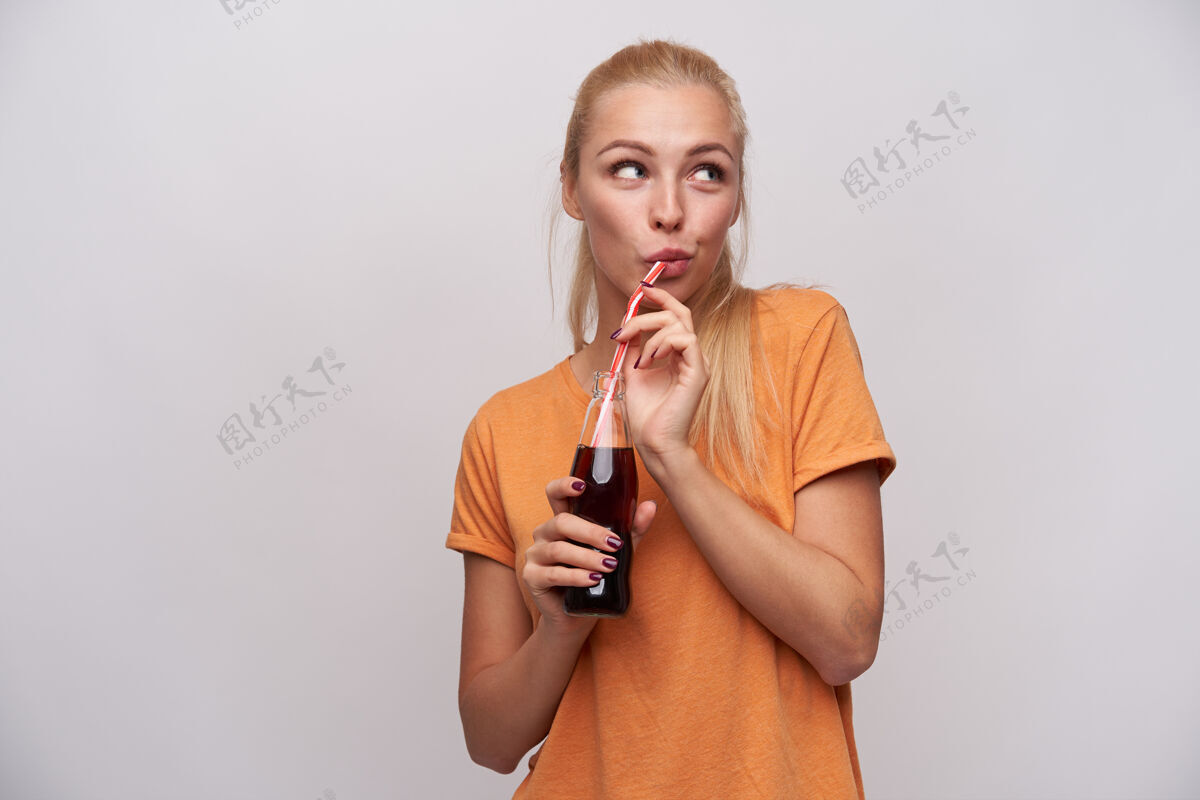 吸管可爱的年轻积极的长发金发女人 随意的发型 喝着玻璃瓶里的苏打水 穿着橙色t恤站在白色背景上年轻手情绪