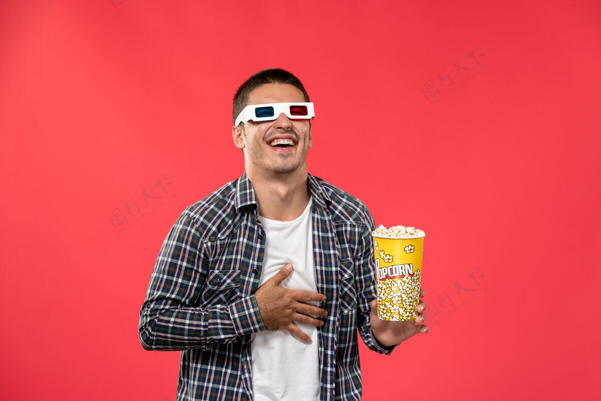 电影正面图年轻男子拿着爆米花包 笑着在淡红色的墙上看电影的时候大笑年轻男人