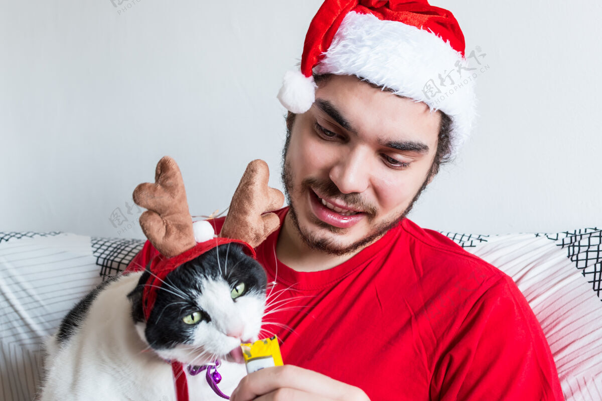 猫一个戴着圣诞老人帽子的白人年轻人 用麋鹿角喂他的黑白相间的猫水平人喂养
