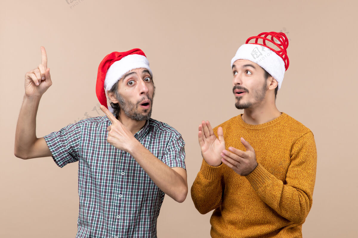 伙计们正面图两个戴着圣诞帽的感兴趣的家伙 一个在米色孤立背景上向另一个展示东西男人帽子男性