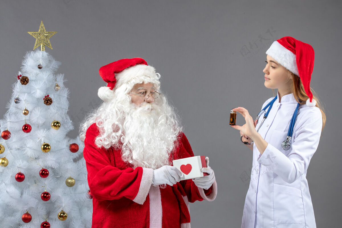 十二月圣诞老人和女医生的前视图 女医生拿着装着药片的小瓶子在灰色的墙上烧瓶圣诞服装