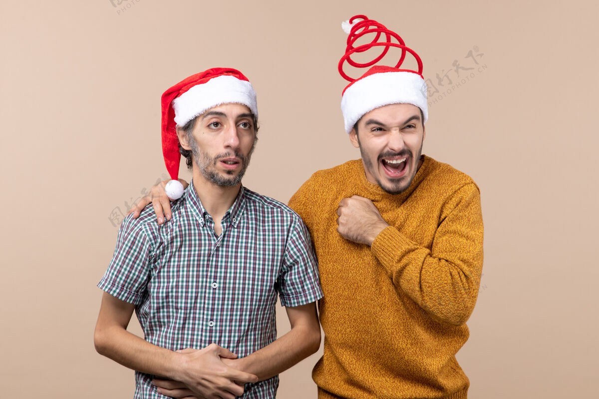 男性正面图两个男人一个沮丧 一个高兴 戴着圣诞帽 一个把手放在其他人的肩膀上 背景是米色的孤立背景帽子两个沮丧