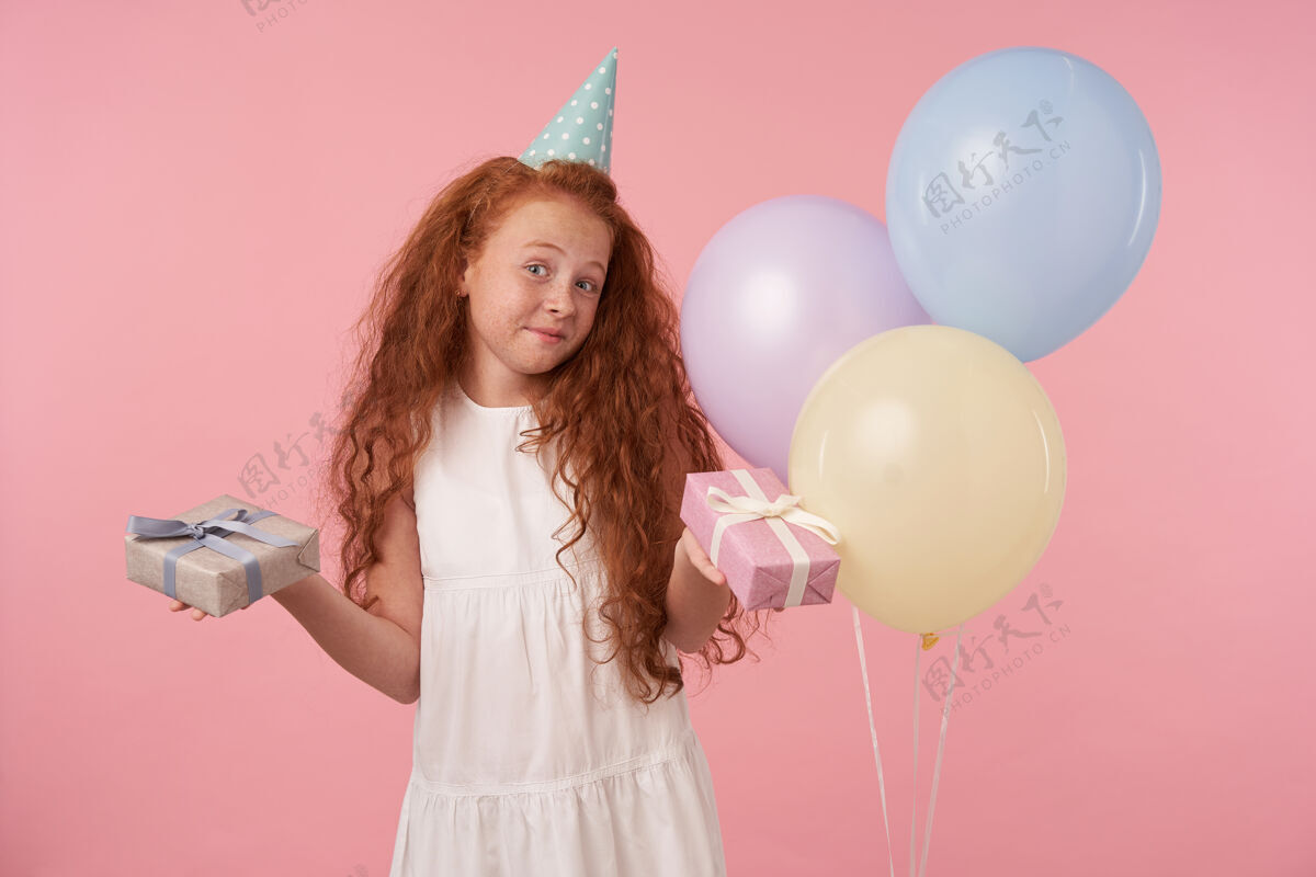 乐趣可爱的小女孩 红色卷发 白色连衣裙 戴着生日帽 高兴地看着镜头 手里拿着礼盒 站在粉色背景和彩色气球的上方礼物卷曲盒子