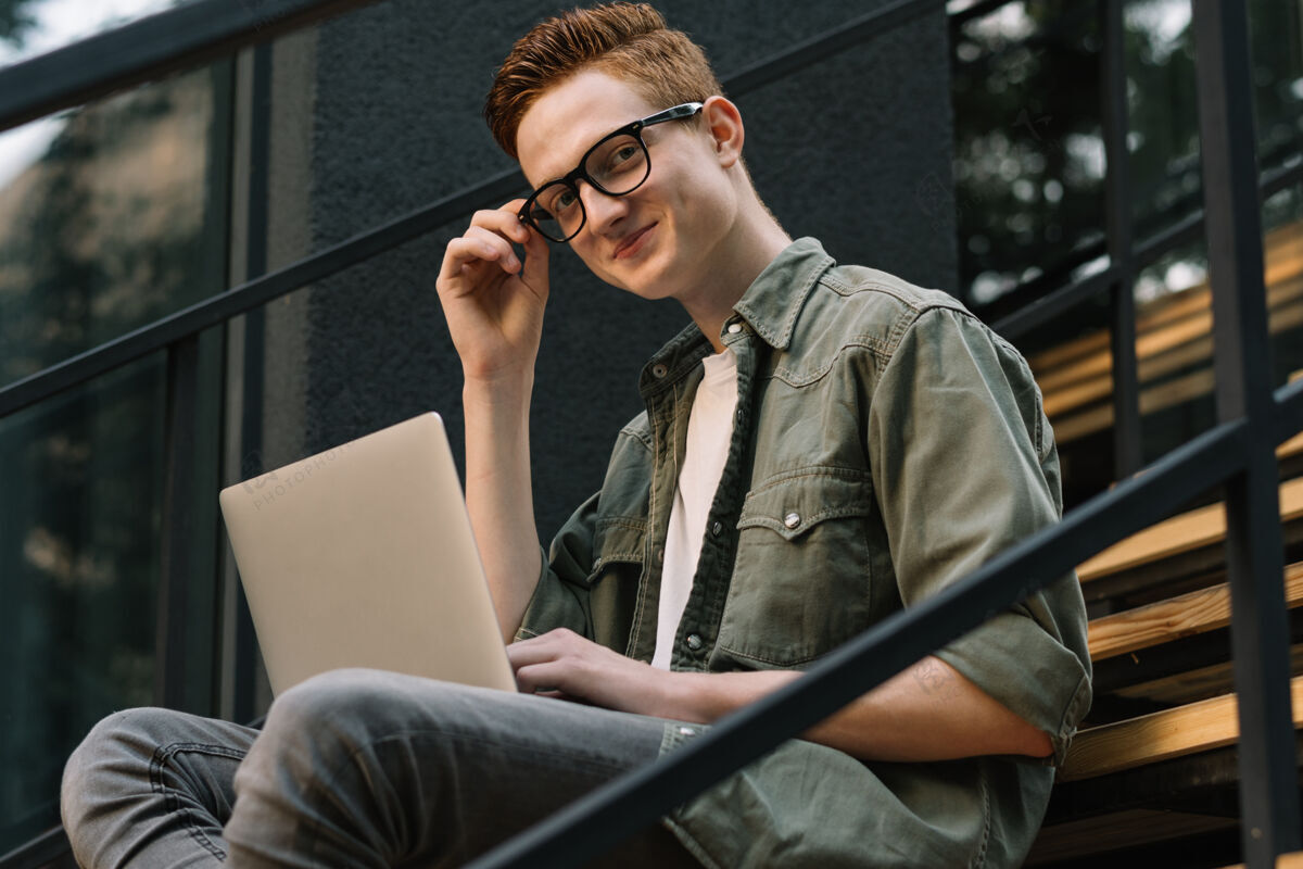 笔记本电脑在街上的楼梯上 一个微笑着用笔记本电脑的年轻人眼镜男人发展