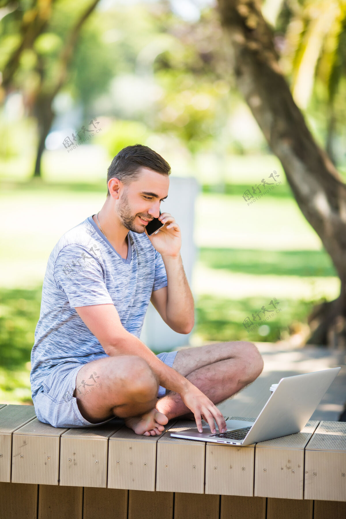 城市一个英俊的年轻人坐在长凳上 拿着笔记本电脑在公园旁边的街道上打电话男士商务公司