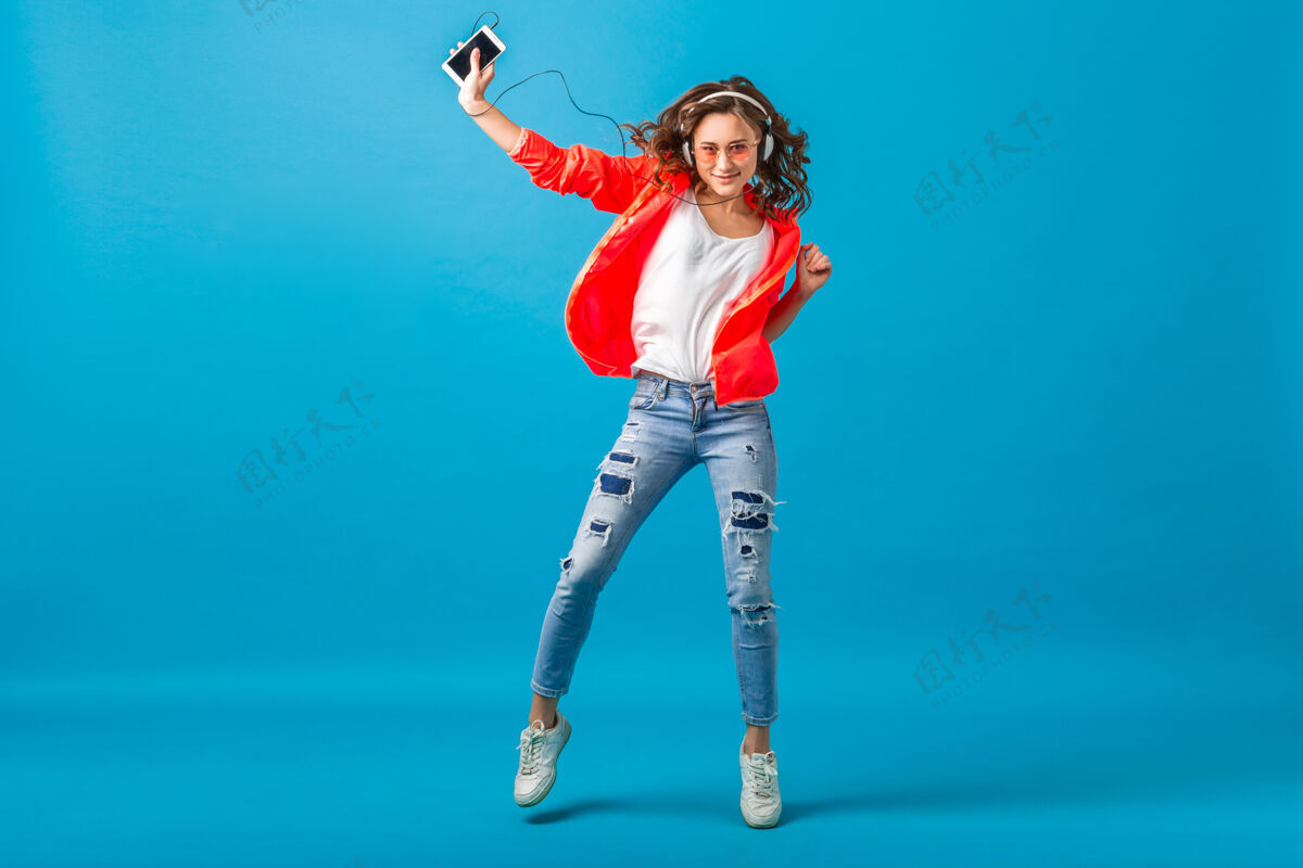 情感迷人的微笑快乐的女人戴着耳机跳舞听音乐穿着时髦风格的服装在蓝色的工作室背景下隔离 穿着粉色夹克和太阳镜听充分五颜六色