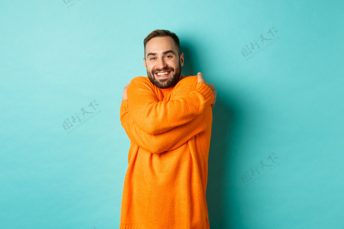 自信快乐的男人感觉很舒服 穿着暖和的毛衣 拥抱着自己 满意地微笑着 站在浅绿的墙上姿势GuyYoung