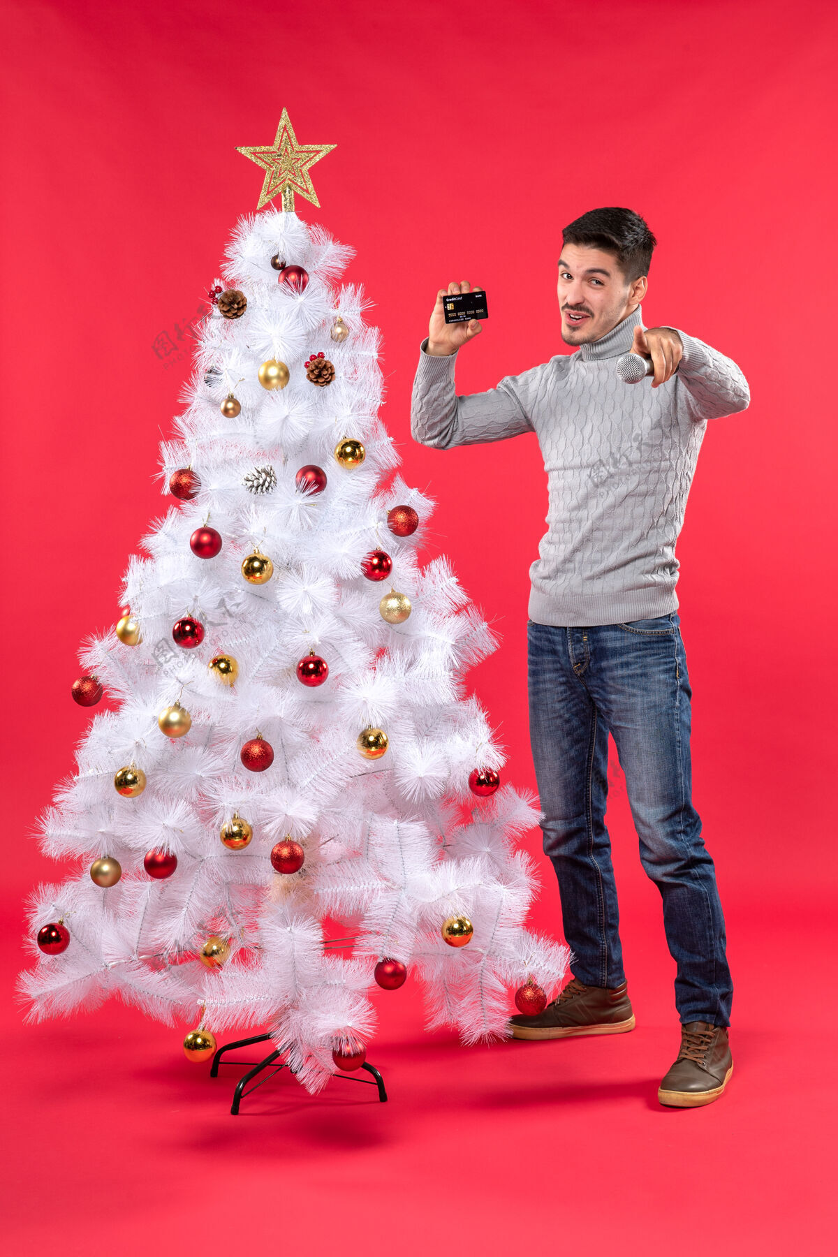微笑圣诞气氛 微笑的年轻人站在装饰好的圣诞树旁 拿着麦克风和电话 指着前方站着的人装饰小伙子抱着
