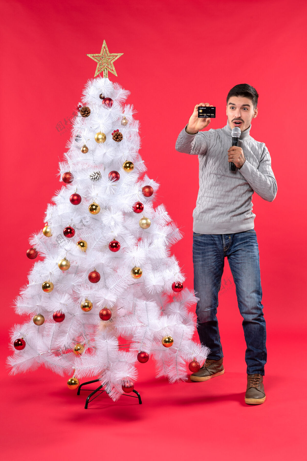 圣诞圣诞节的气氛 自信的家伙穿着牛仔裤站在装饰圣诞树附近 拿着麦克风拍照自信站着微笑