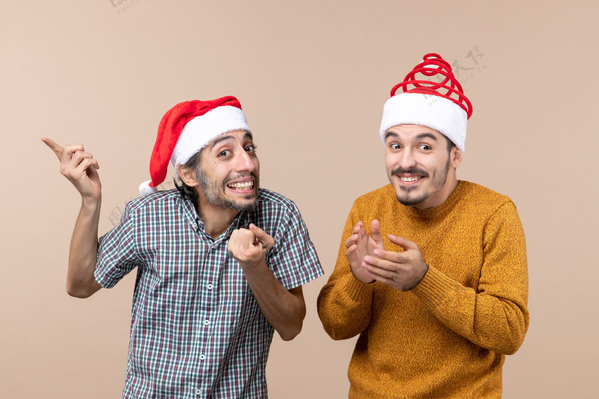 两个前视图两个带着圣诞帽的微笑的家伙一个在米色孤立背景上展示东西伙计们帽子肖像