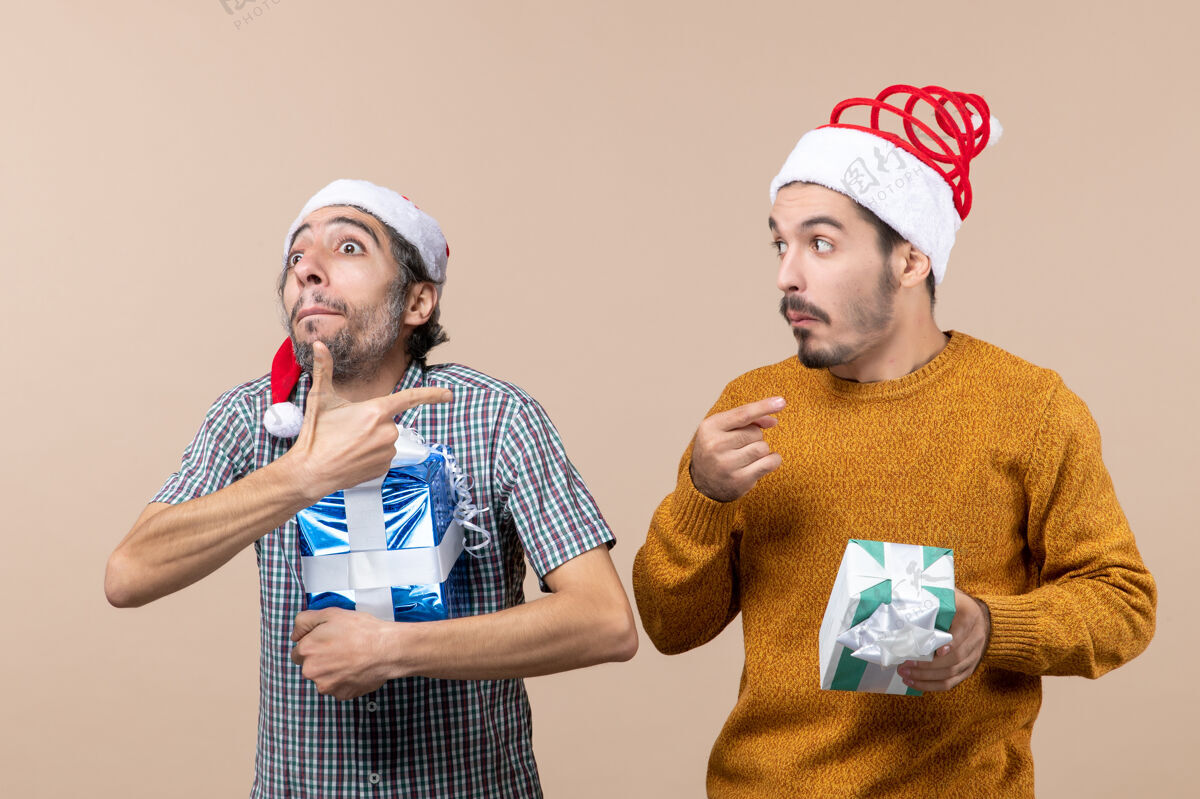 两个前视图两个困惑的家伙手指展示东西 拿着圣诞礼物在米色孤立的背景肖像两个困惑的家伙持有