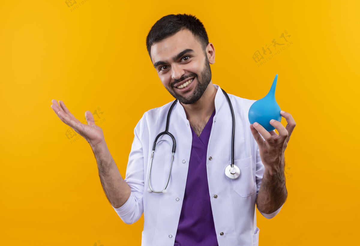 微笑微笑的年轻男医生穿着听诊器医用长袍 在孤立的黄色背景下拿着灌肠器抱着穿着男性