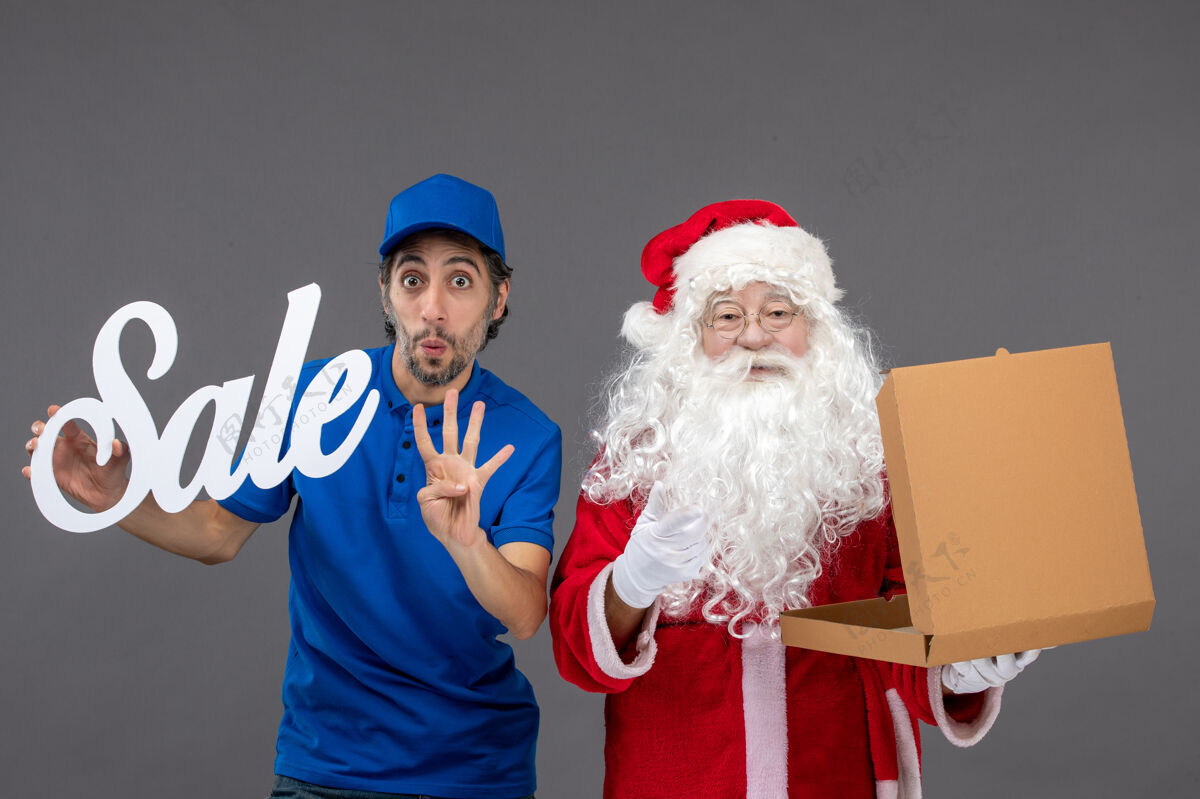 人圣诞老人的正面图 男信使拿着卖东西的字条和灰墙上的食品盒快乐圣诞老人服装