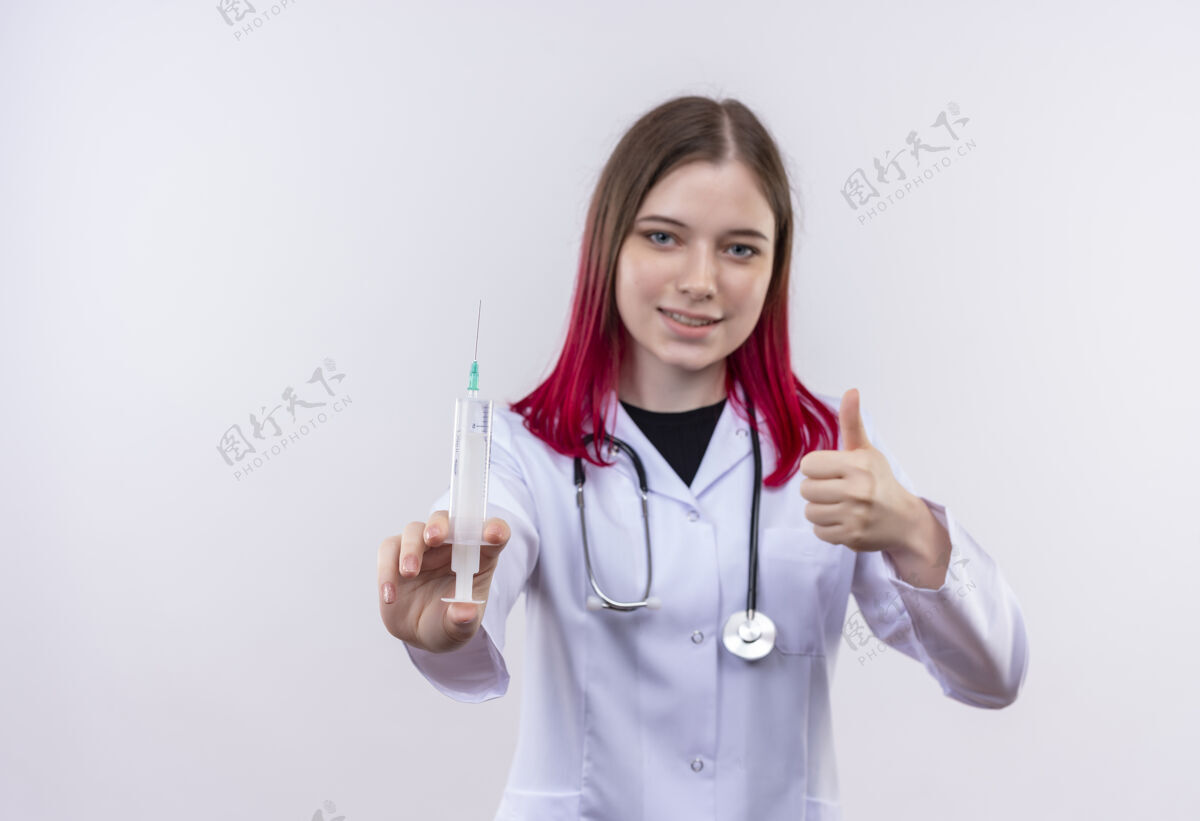 听诊器高兴的年轻医生女孩穿着听诊器医用长袍拿着注射器她的拇指上孤立的白色背景拇指白色向上
