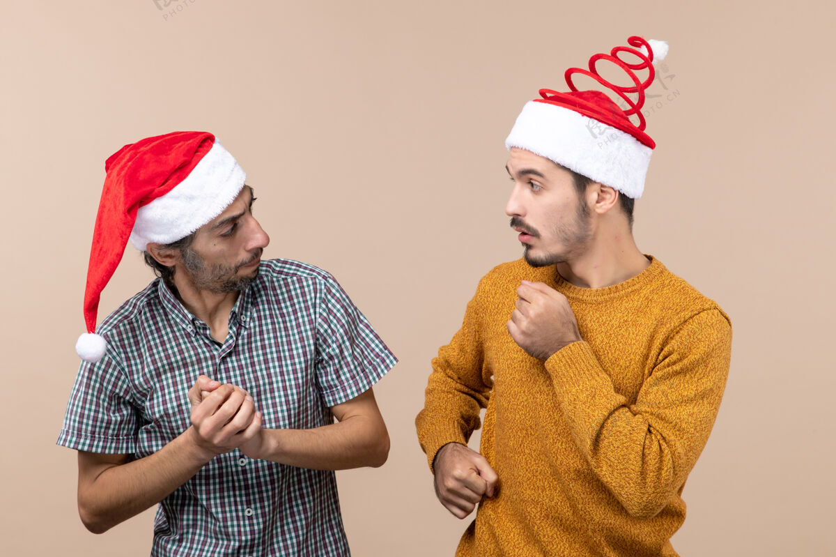 男前视图两个圣诞老人戴着圣诞帽的家伙在米色孤立的背景下饶有兴趣地看着对方男人帽子圣诞老人
