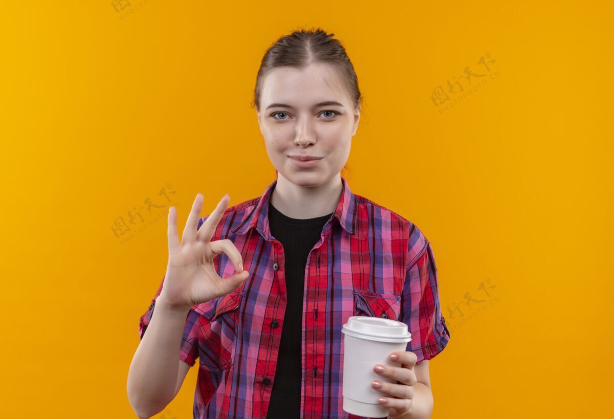 空间高兴的年轻漂亮的女孩穿着红衬衫 拿着一杯咖啡 在孤立的黄色背景和复制空间上显示okey手势穿着高兴杯子