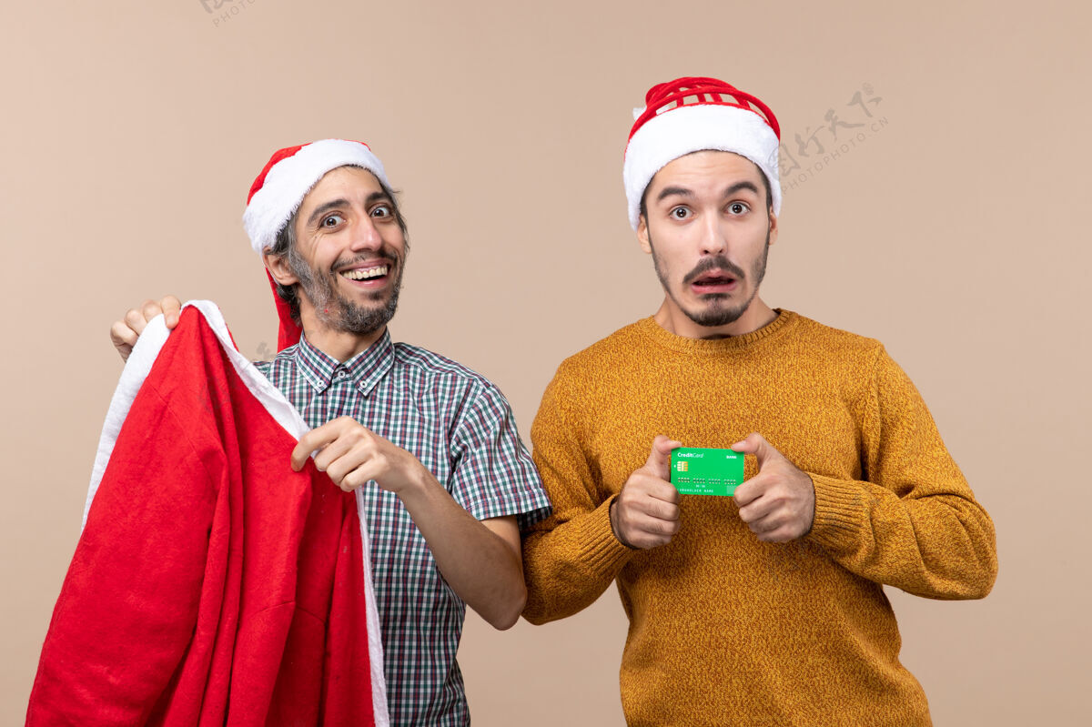 视图前视图两个家伙一个拿着圣诞老人的外套 另一个拿着信用卡 在米色孤立的背景下看着摄像机微笑圣诞老人看