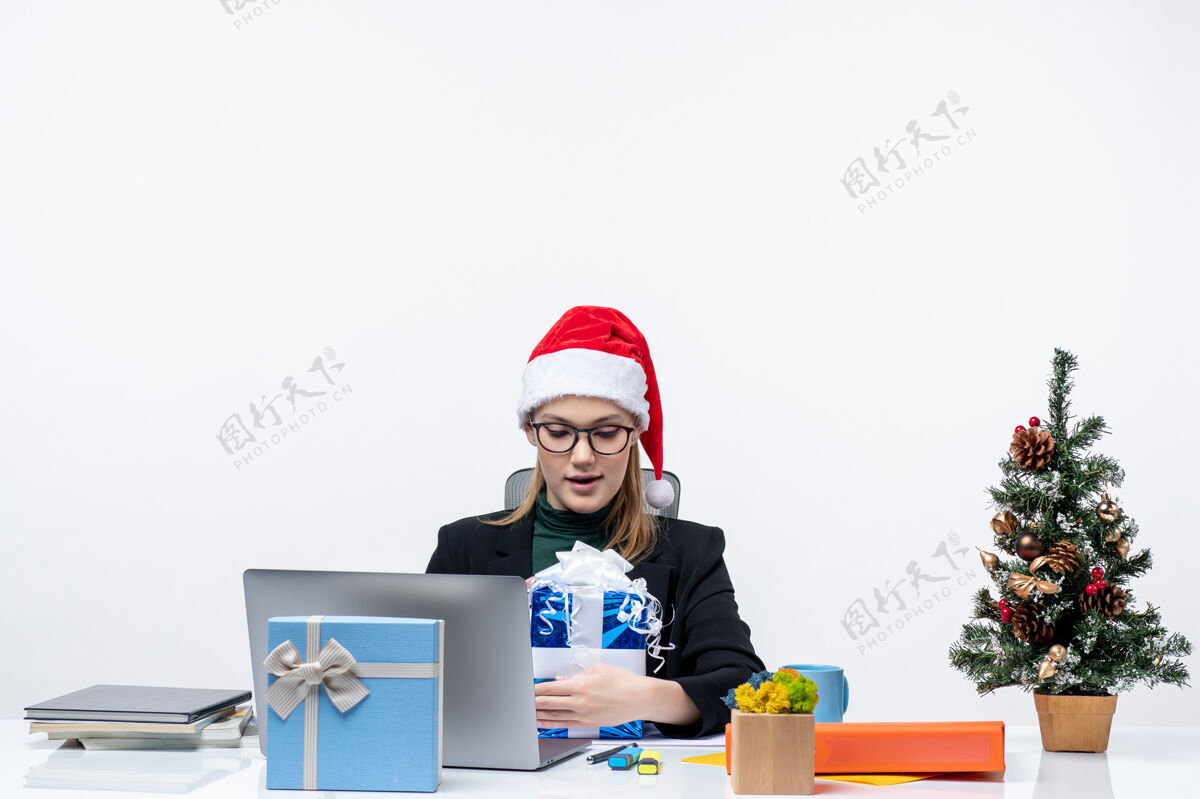 圣诞老人圣诞快乐的年轻女子戴着圣诞老人的帽子 戴着眼镜 坐在桌子旁骄傲地看着白色背景上的礼物电脑年轻人桌子