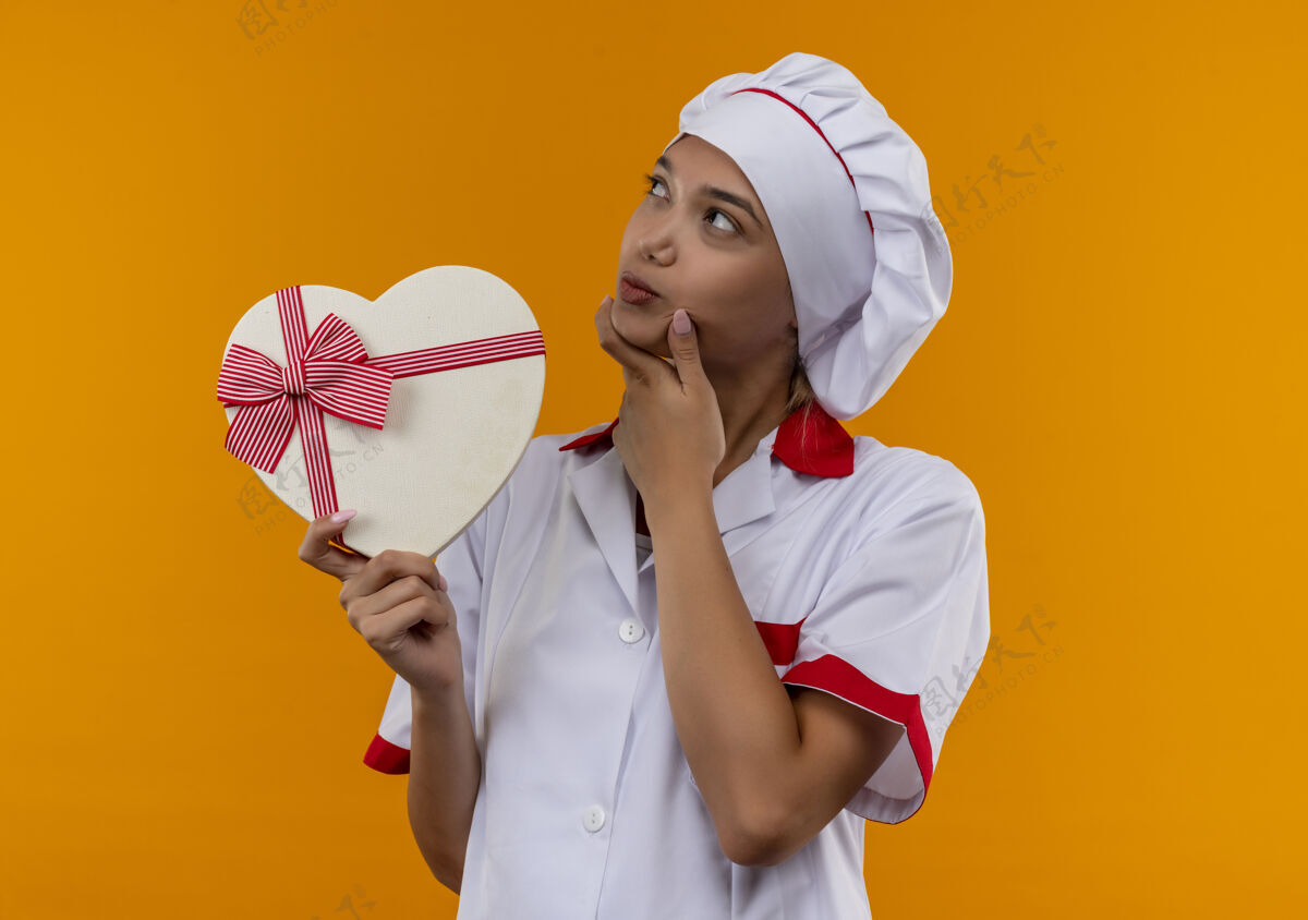 厨师想着年轻的女厨师穿着厨师制服拿着心形盒子把手放在下巴上孤立的橙色背景戴着厨师制服