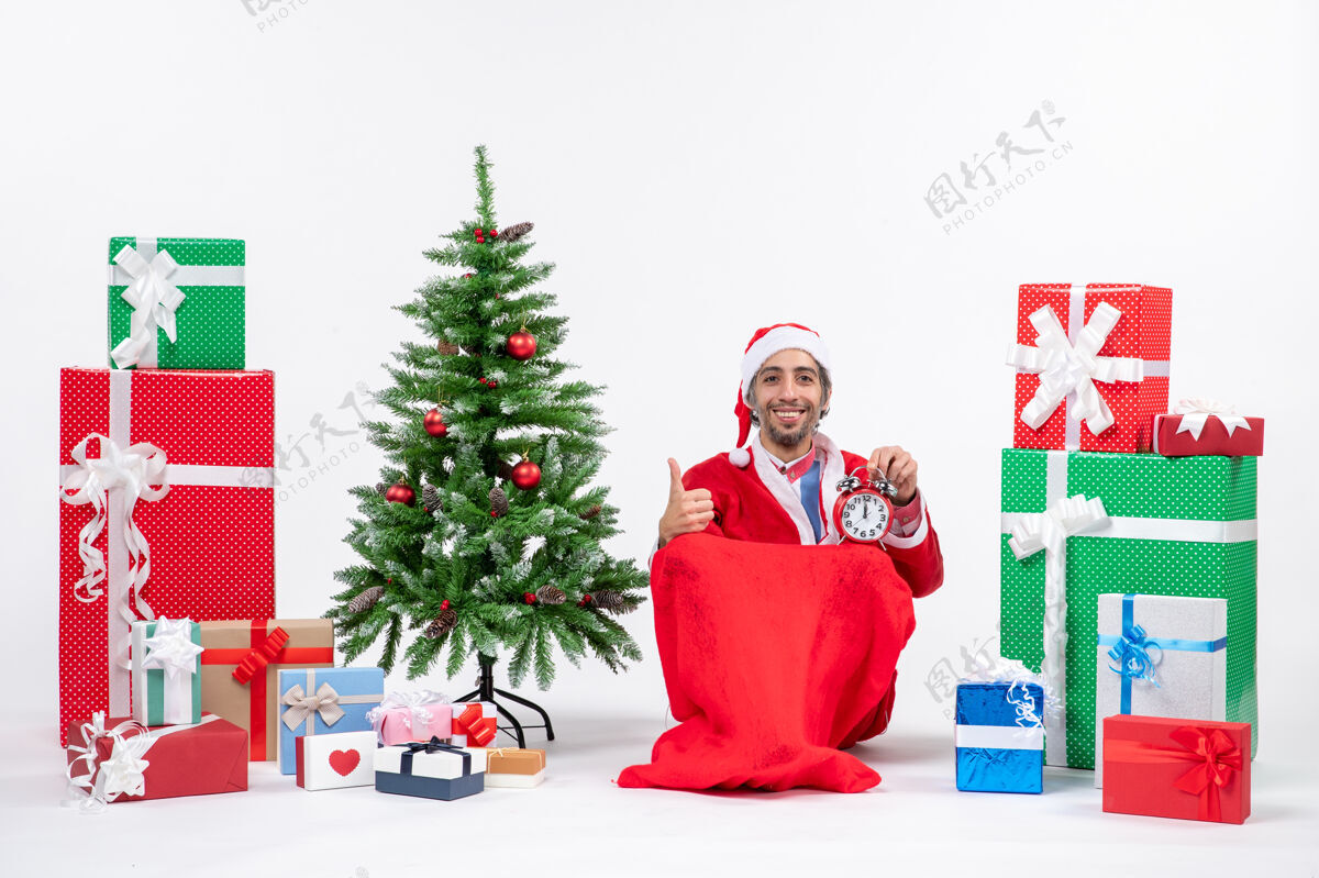 礼物微笑的年轻人坐在地上 拿着钟 在礼物和装饰过的圣诞树旁庆祝新年或圣诞节年轻人抱着男人