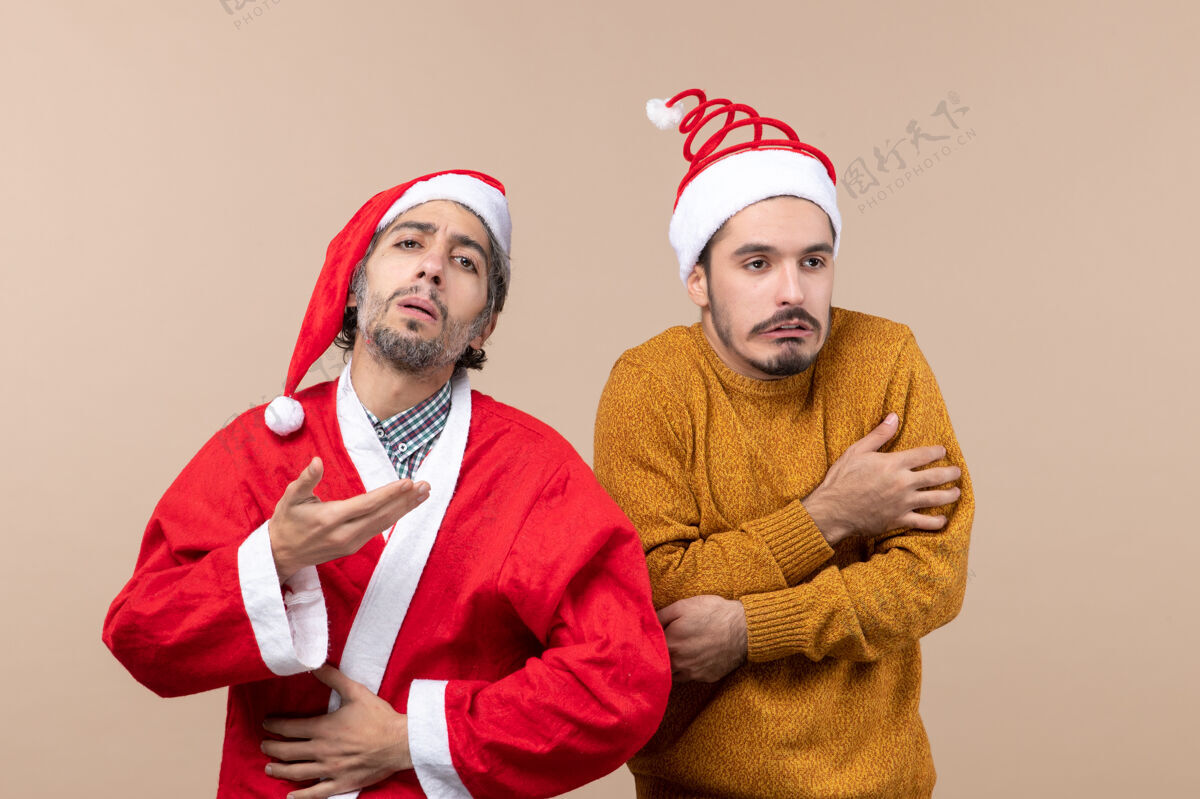 伙计们前视图两个圣诞节的家伙一个穿着圣诞老人外套 但另一个冻结在米色孤立的背景外套圣诞老人帽子