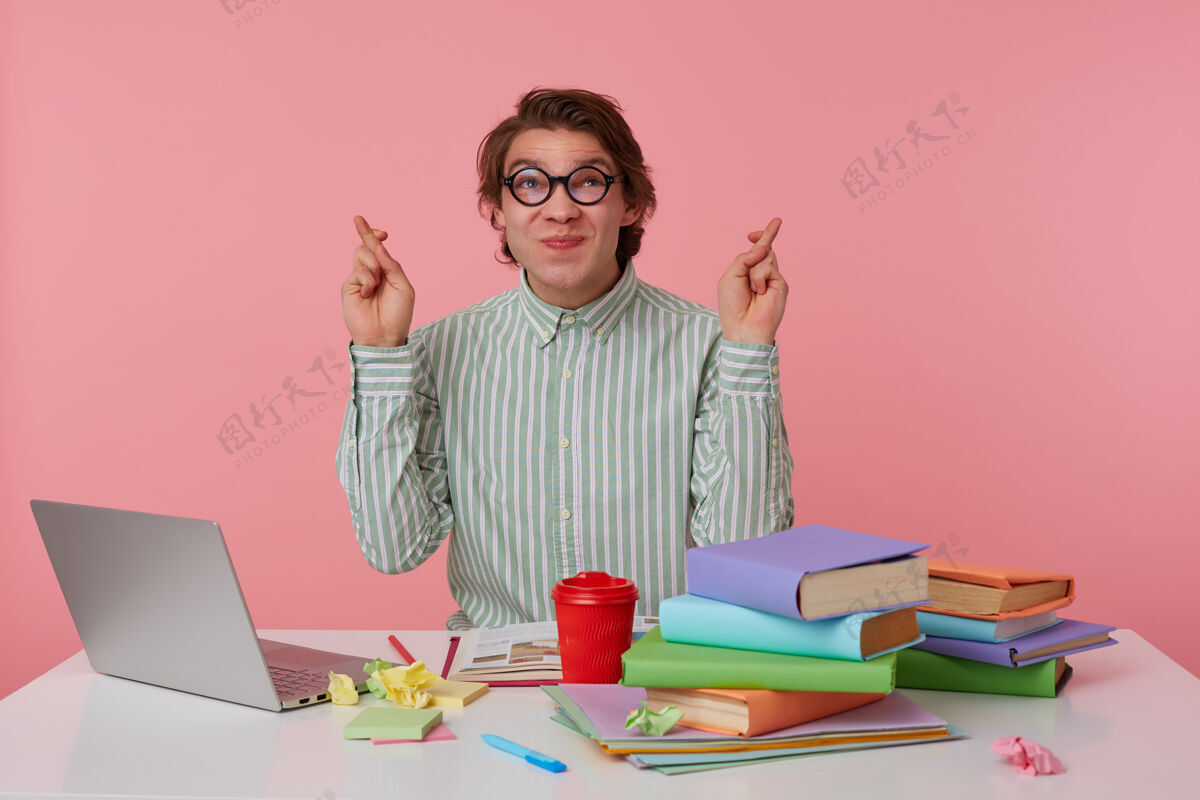 男性戴着眼镜的快乐的年轻黑发男人举起交叉的手指许愿 穿着条纹衬衫在工作台摆姿势准备坐着笔记本