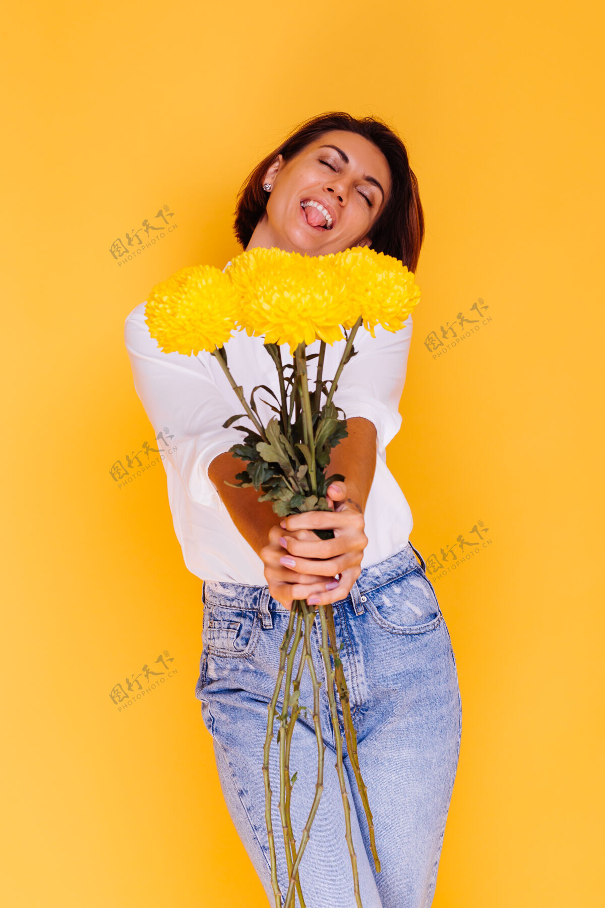 植物摄影棚拍摄的黄色背景快乐的白人妇女短发穿着休闲服白衬衫和牛仔裤手持一束黄色紫苑积极礼物年轻