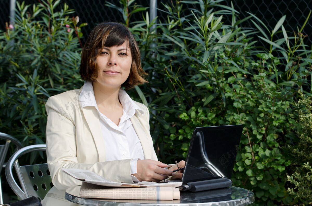 经理一个穿着白色西装的女人坐在一张桌子旁 她的笔记本电脑靠近绿色的灌木丛流程互联网电话