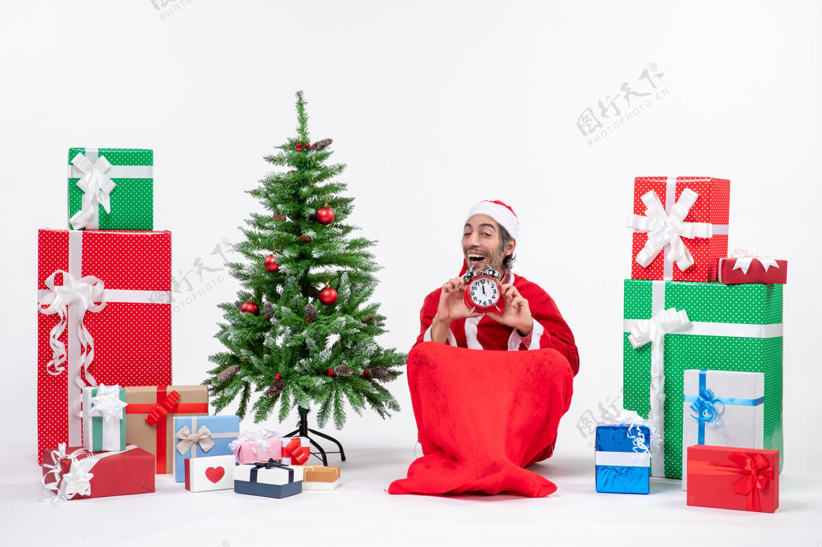 坐微笑快乐满足的年轻人庆祝新年或圣诞节假期坐在地上 拿着礼物和装饰圣诞树附近的时钟圣诞节礼物装饰