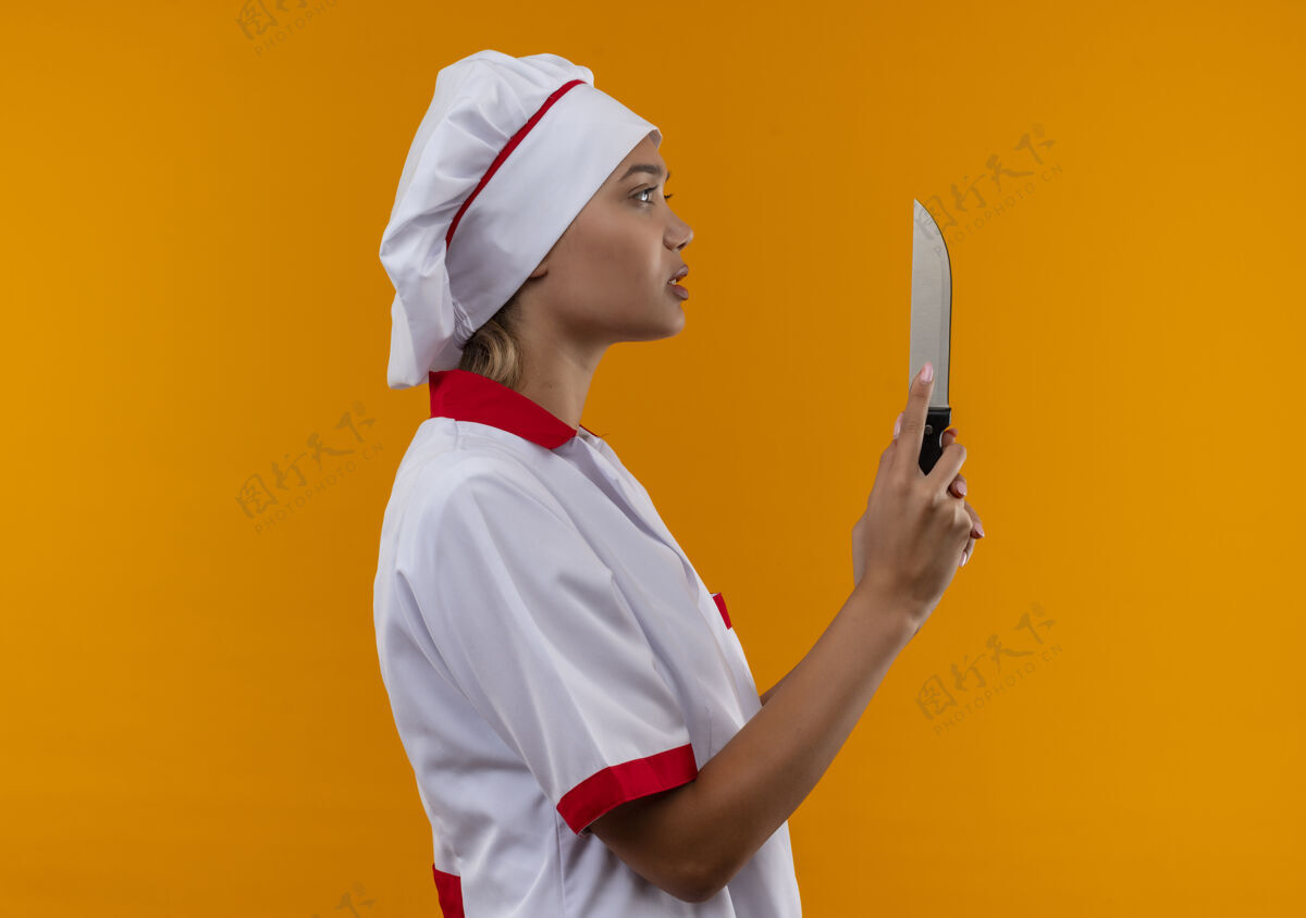 女站在剖面图年轻厨师女身穿厨师制服持刀在孤立的橙色背景与复制空间刀厨师拿着