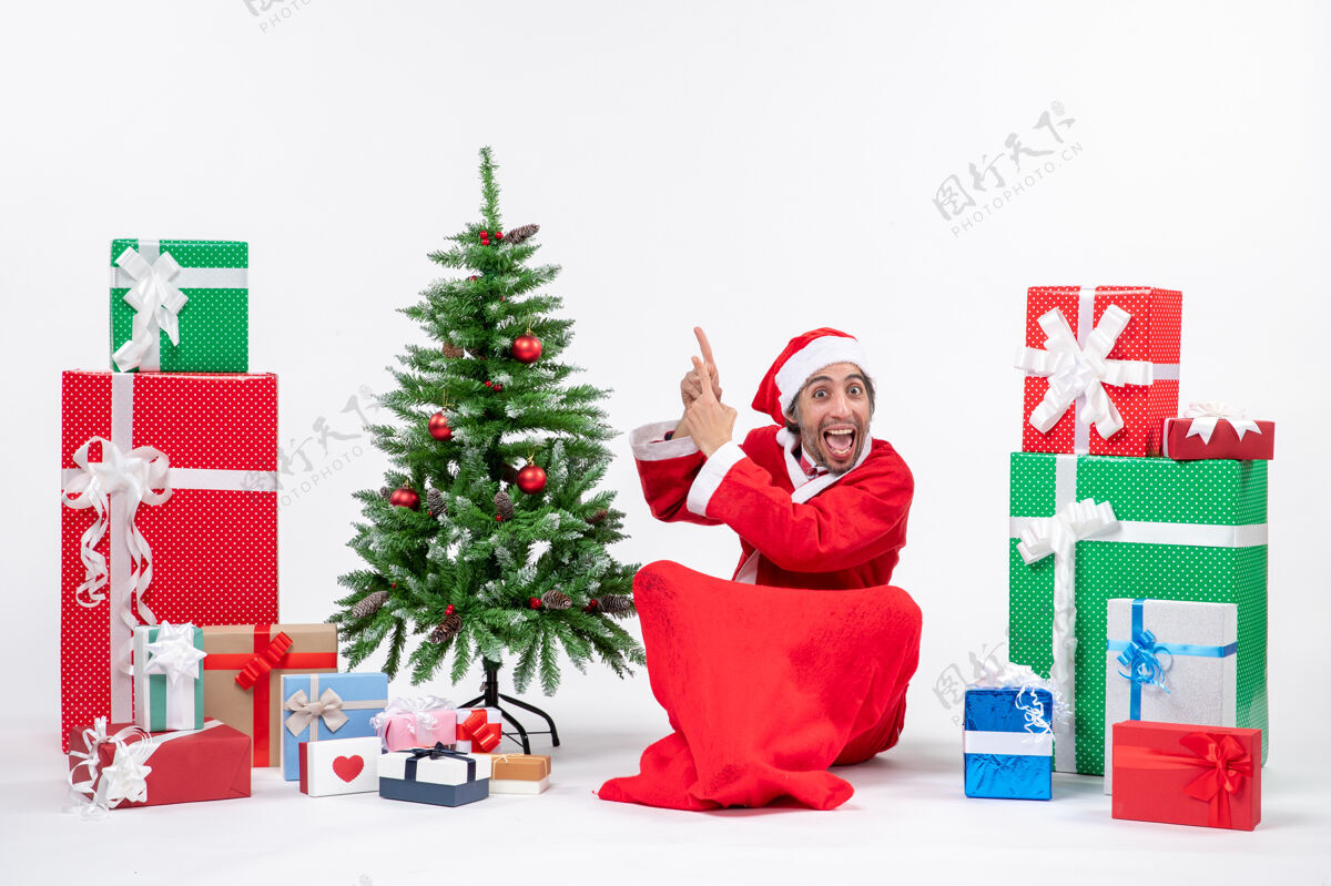 礼物有趣的年轻人打扮成圣诞老人与礼物和装饰圣诞树坐在地上指着上面的白色背景圣诞圣诞树地面