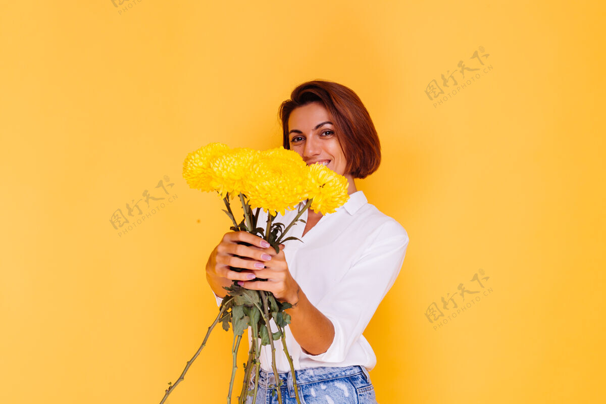积极摄影棚拍摄的黄色背景快乐的白人妇女短发穿着休闲服白衬衫和牛仔裤手持一束黄色紫苑年轻植物惊喜
