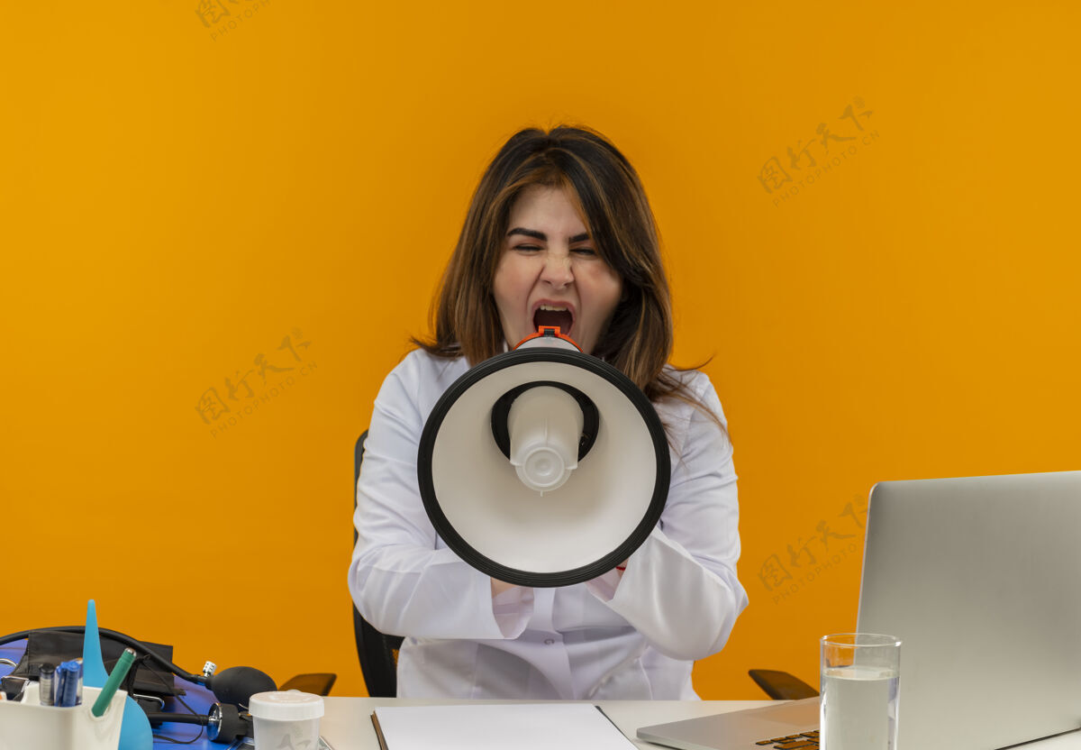 穿着闭着眼睛 穿着医用长袍 带听诊器的中年女医生坐在办公桌旁 用笔记本电脑和医疗工具 在隔离的橙色背景和复印空间上用扬声器讲话医疗坐着工作