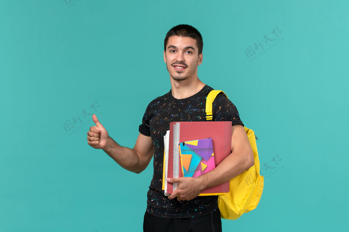 快乐身穿深色t恤 背着黄色背包 拿着文案和文件的男学生的正面图 蓝色墙上书学院成人