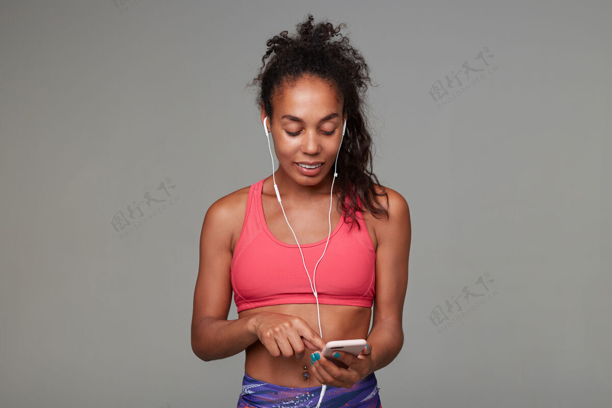 倾听积极的年轻黑皮肤卷曲的黑发女运动员 马尾辫发型 举着手机 愉快地微笑着 身着运动粉色上衣衣服形状女性