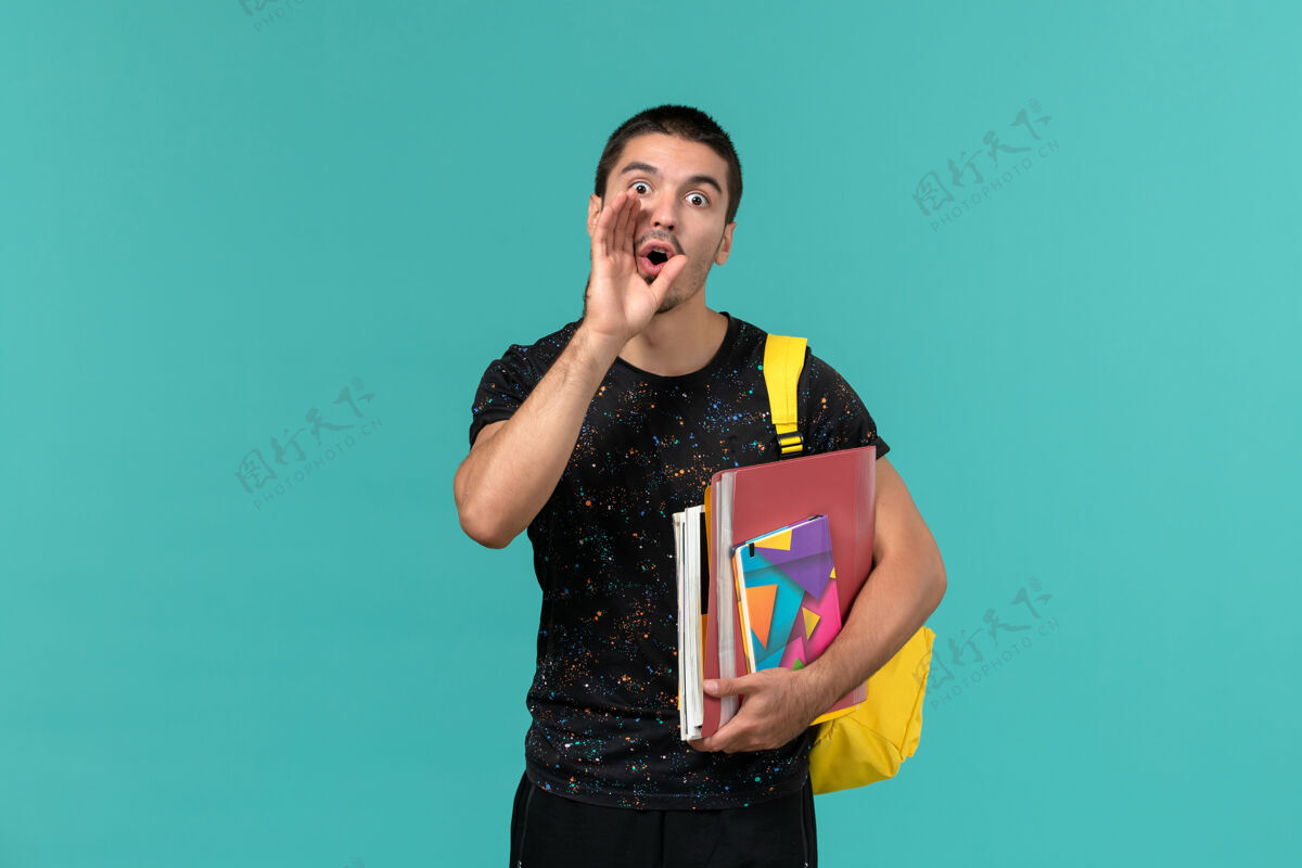 书身穿深色t恤 背着黄色背包 手里拿着抄写本和文件的男学生正对着蓝色墙壁大声呼喊背包学生穿着