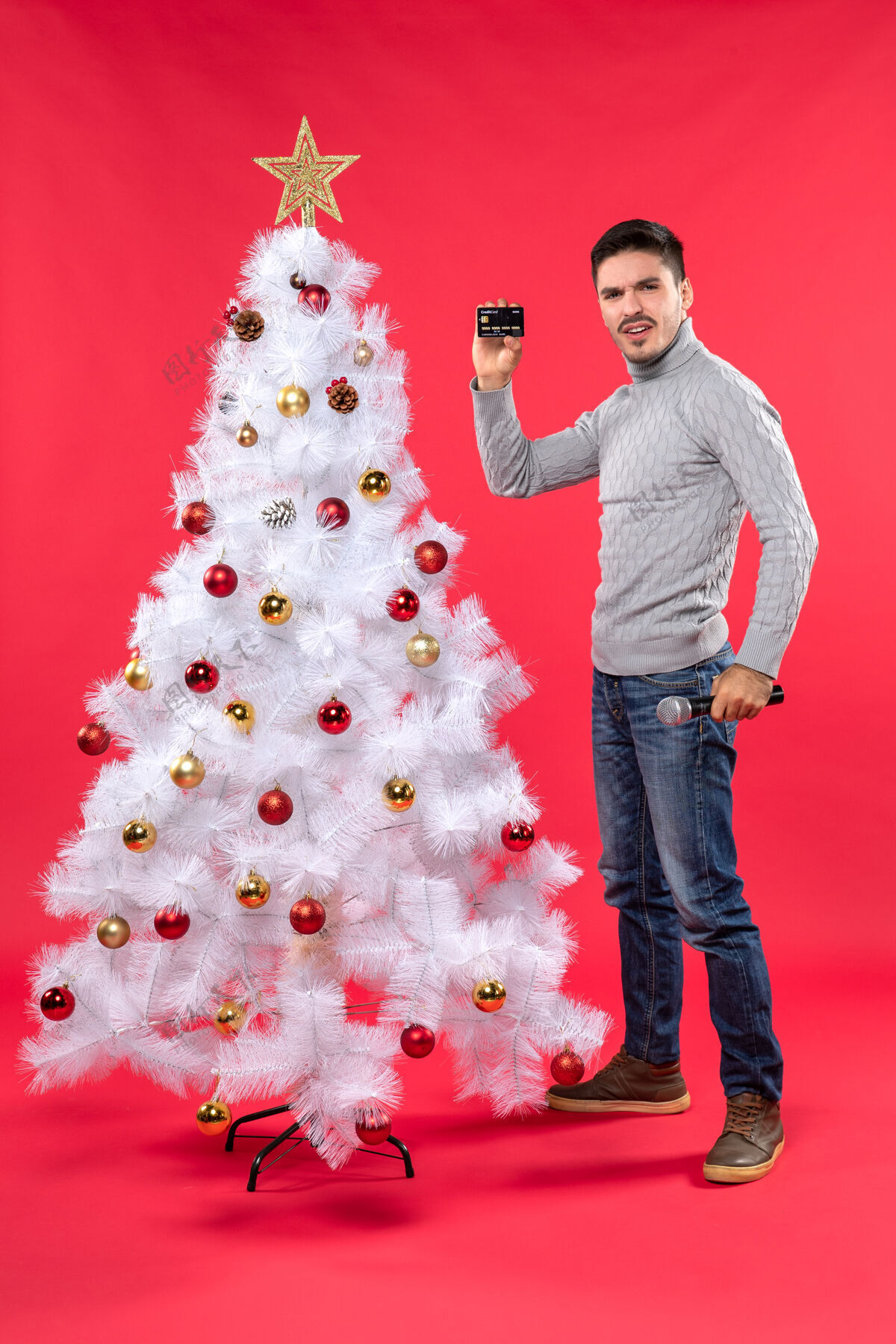 年轻圣诞气氛严肃的年轻人站在装饰好的圣诞树旁 拿着麦克风和电话自信地站着站着季节装饰