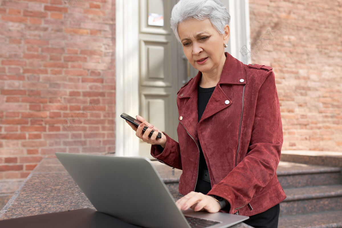 通讯户外拍摄的严肃的灰色头发的女房地产经纪人穿着时尚的衣服站在砖房外打开笔记本电脑前 使用无线互联网连接女性连接工作