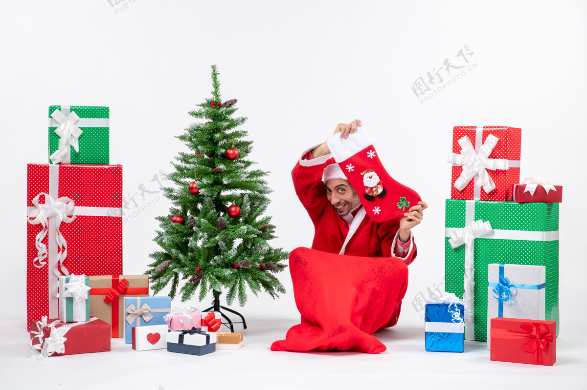 惊讶喜庆的节日气氛 有趣的积极惊喜的圣诞老人坐在地上 展示圣诞袜子附近的礼物和装饰圣诞树上的白色背景礼物礼物地面