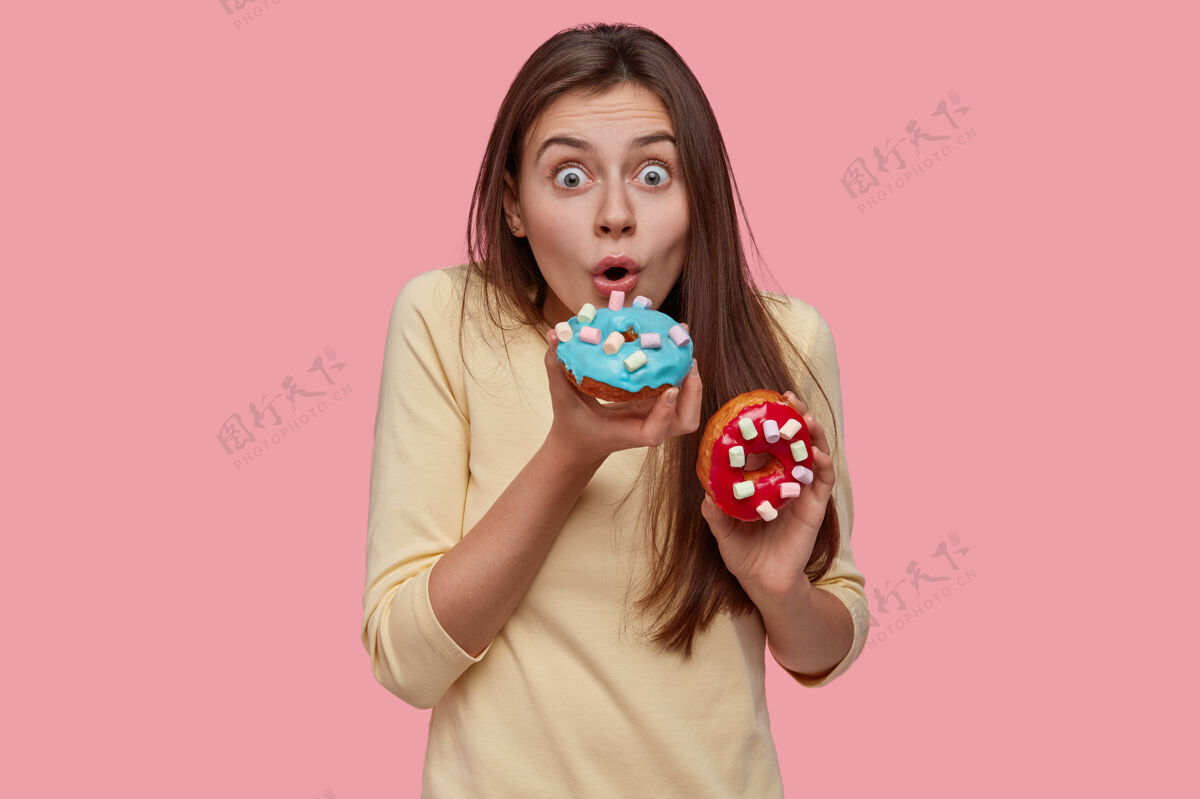 人类惊讶的年轻可爱的欧洲女人伯特斯闪耀美味的甜甜圈 睁大眼睛 穿着黄色毛衣 有美味的小吃天哪食物节食