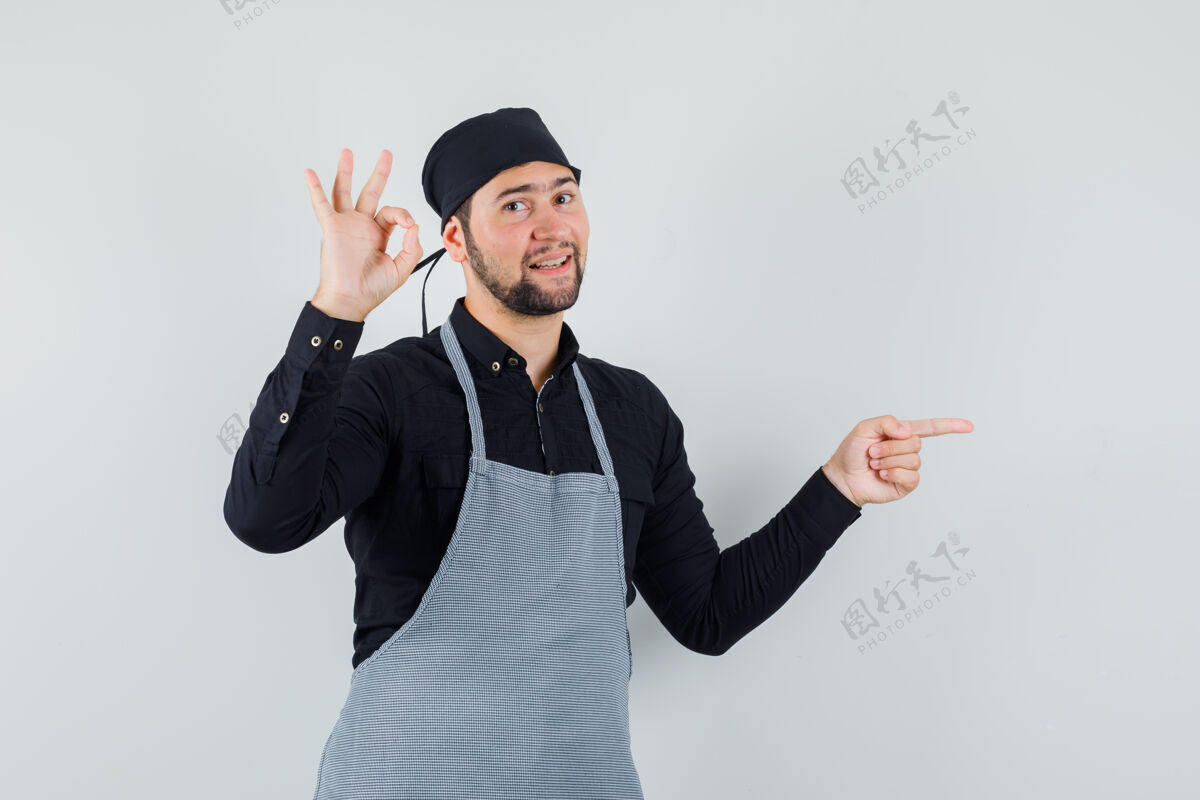 厨师男厨师展示ok标志 同时指着衬衫 围裙 看起来很高兴 正面视图美味手烹饪