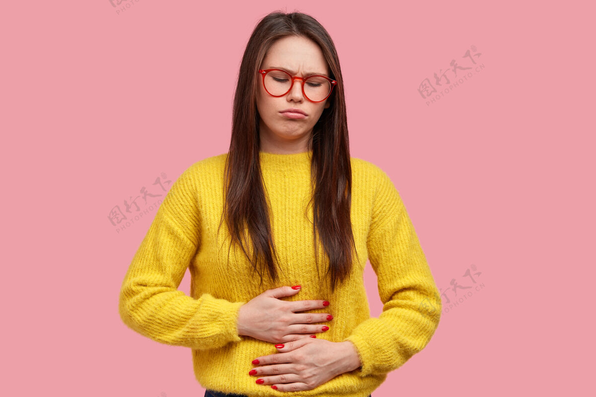不满漂亮女人把手按在肚子上 感到疼痛和不适 有健康问题和饮食不适 穿着黄色毛衣 在粉色背景下摆姿势疾病毛衣经期