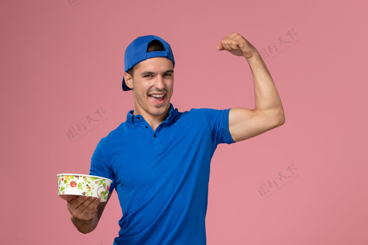 人正面图：身穿蓝色制服披肩的年轻男性信使手持圆形投递碗 在浅粉色的墙上弯腰送货粉红制服