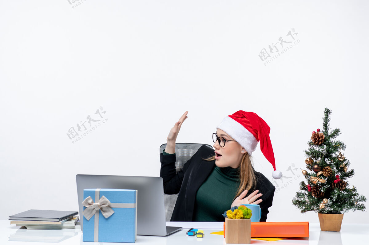 圣诞树一个戴着圣诞老人帽子的年轻女子坐在一张桌子旁 桌子上放着圣诞树和礼物 手指着白色背景坐着笔记本电脑年轻女人
