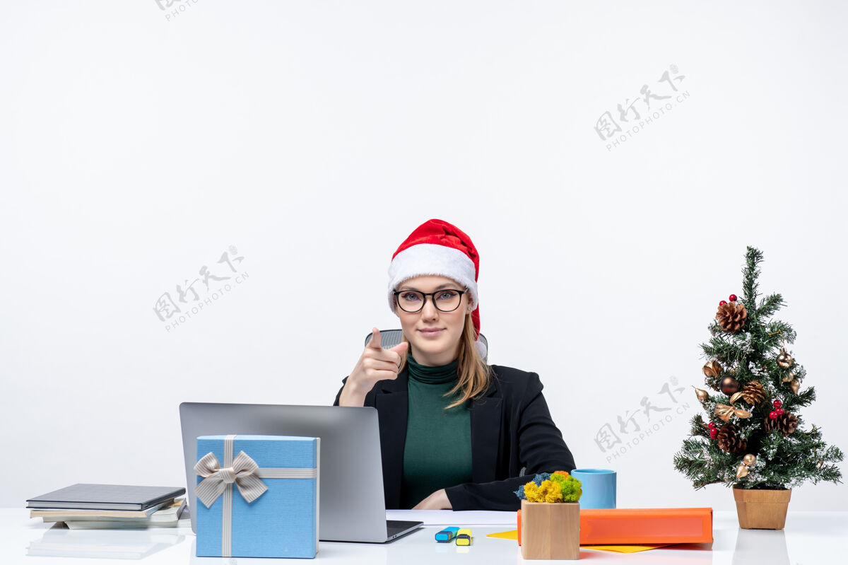 金发女人新年的气氛与金发女人坐在一张桌子上 圣诞树和一个白色背景的礼物圣诞老人帽子女人圣诞树办公室
