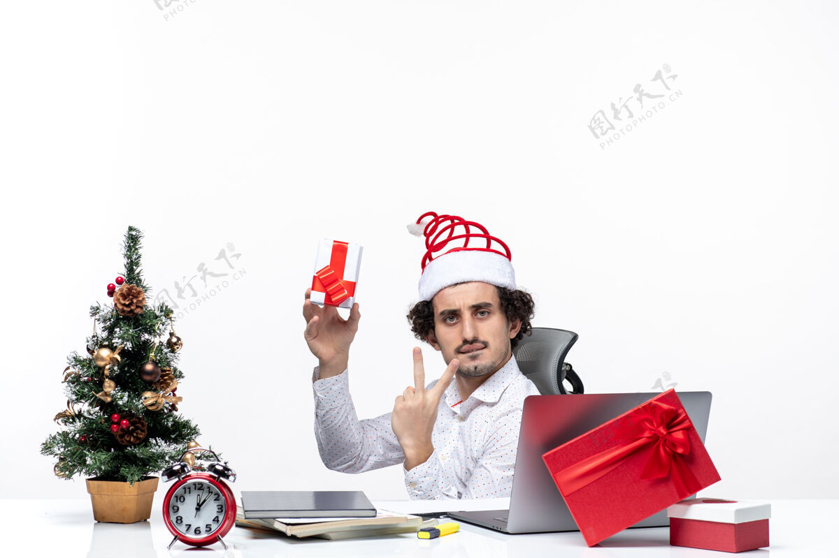 微笑新年气氛与年轻的商人与圣诞老人的帽子坐在办公室里 拿着他的礼物显示两个白色背景抱着坐着礼物