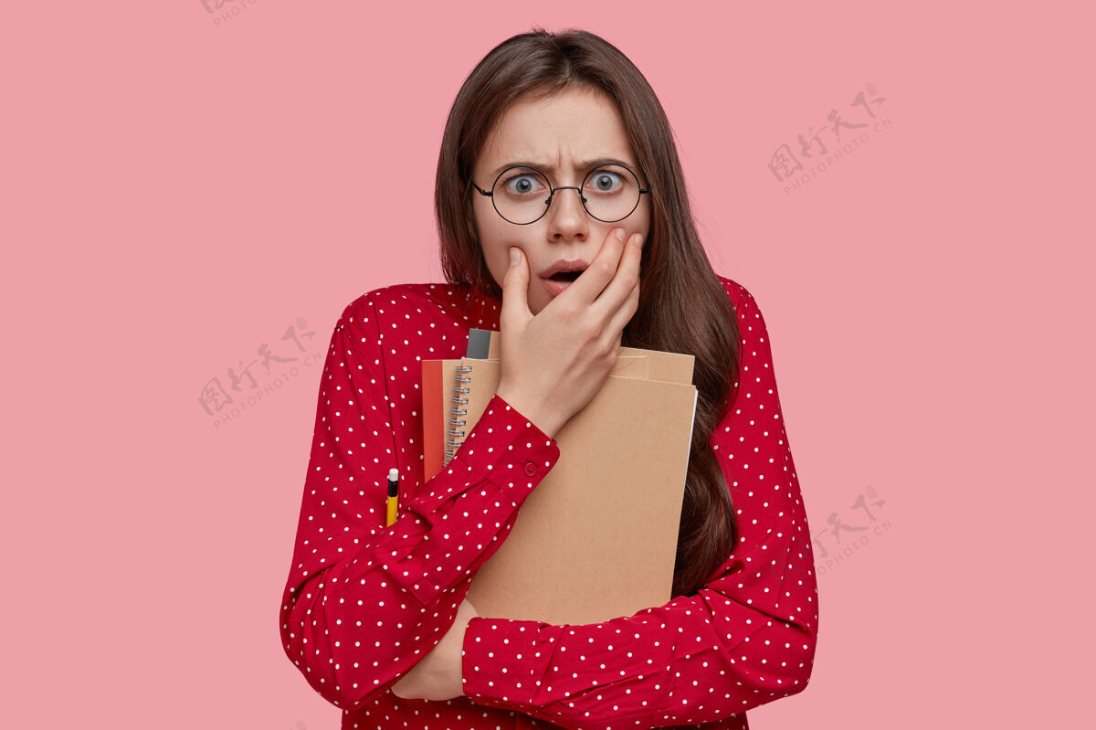 害怕害怕的少年皱着眉头 戴着眼镜 拿着铅笔的记事本 穿着红衬衫 模特们站在粉色的工作室墙上铅笔体贴考试