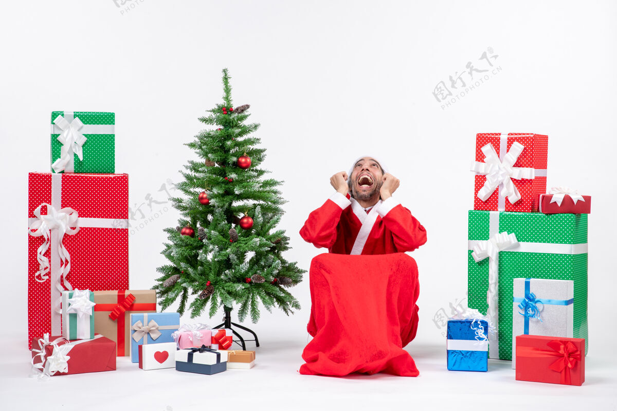 情绪化情绪激动的关心的年轻人打扮成圣诞老人与礼物和装饰圣诞树上的白色背景打扮微笑微笑