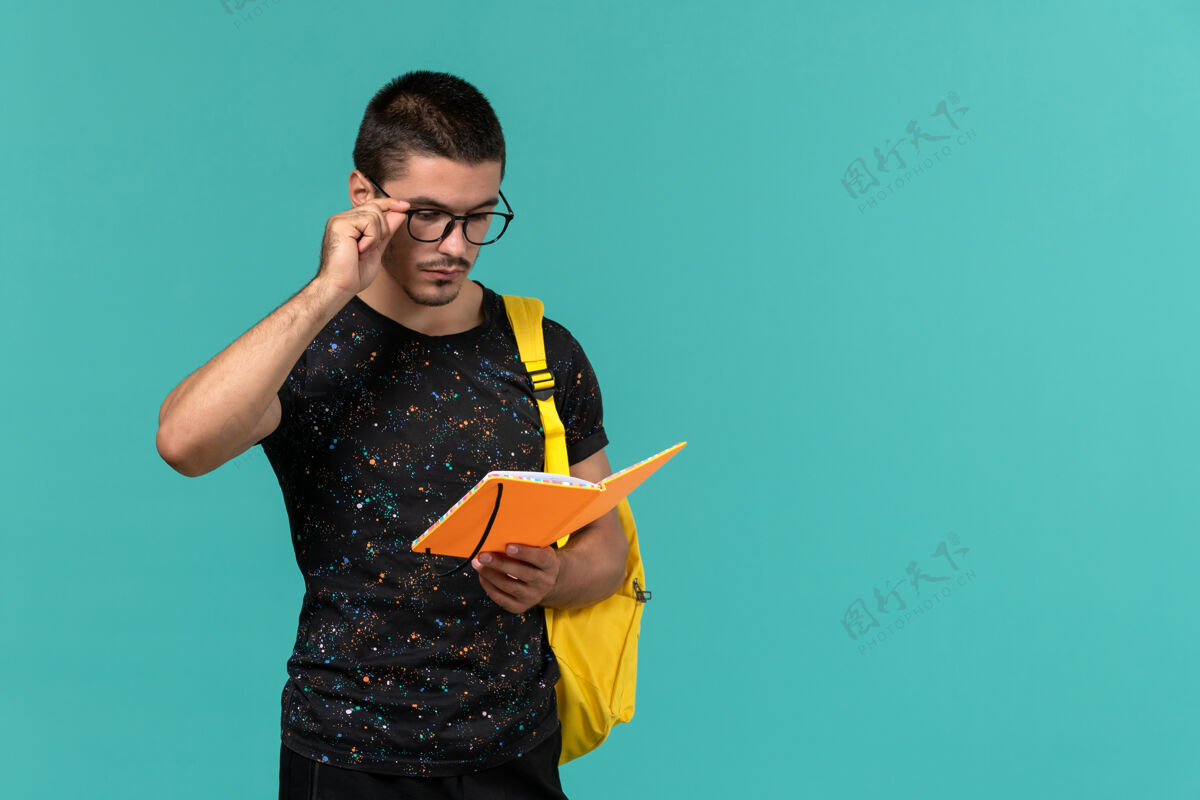 书身穿深色t恤黄色背包的男学生的正面图拿着蓝色墙壁上的抄写本阅读教训学生模型