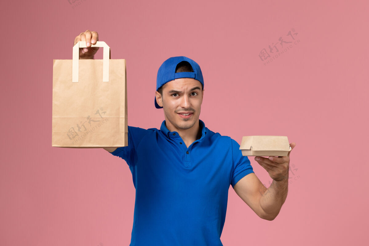 年轻正面图：身穿蓝色制服和披风的年轻男性快递员 手上拿着快递包裹 站在粉红色的墙上视野送货工作